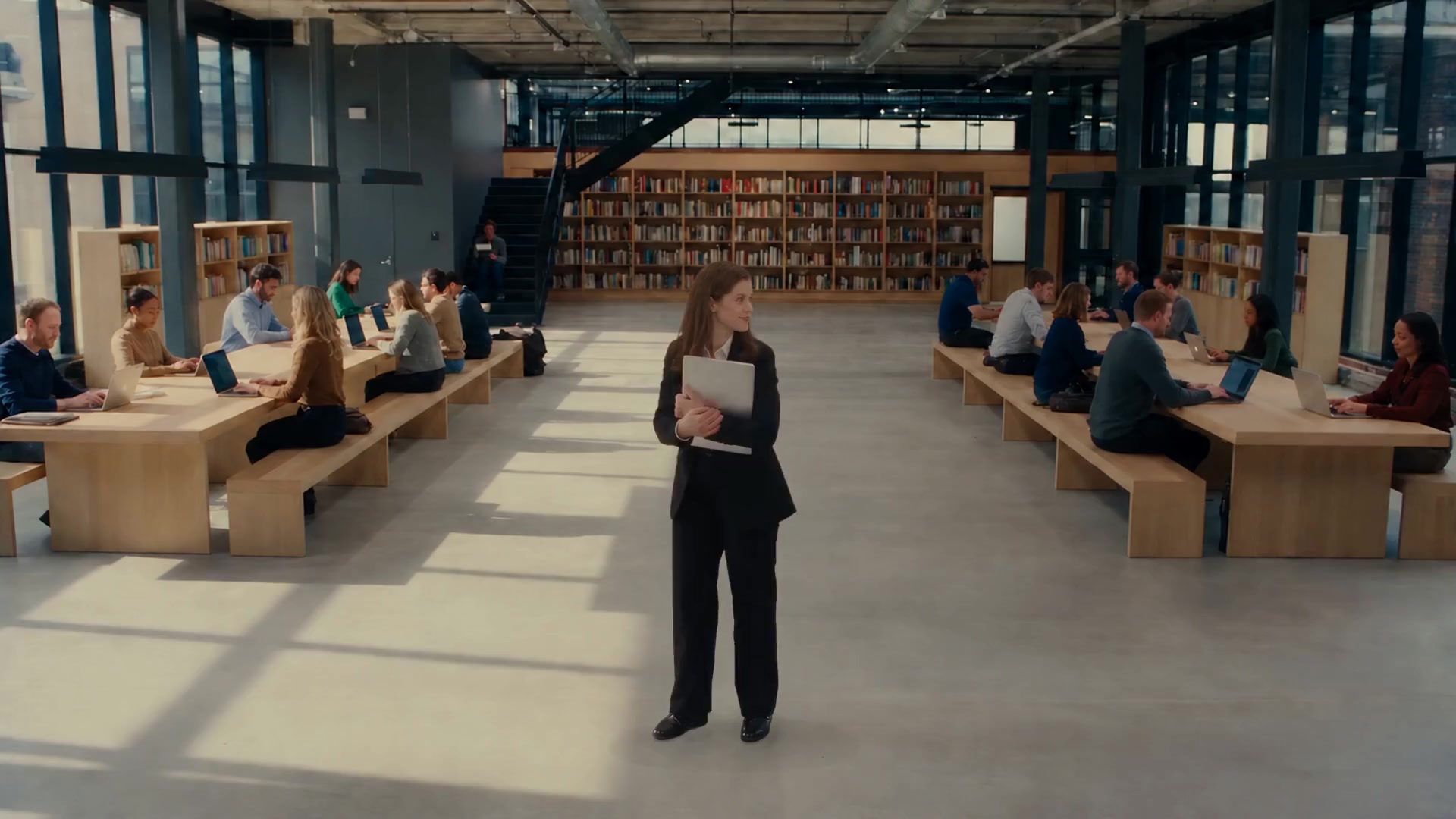 A woman with a laptop smiles as she crosses a bright open-plan office, then tries a bench, a lounge chair, the kitchen and the stairs, each friendly and busy, none of them quiet enough to work