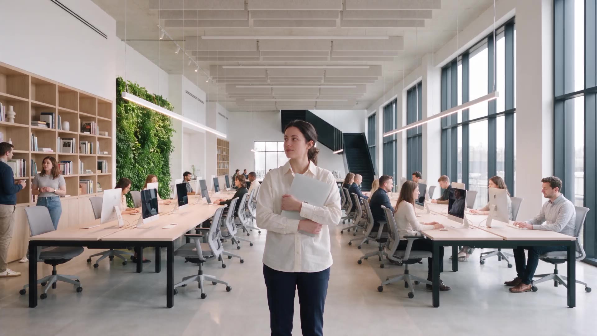 A woman stands still with her laptop in a bright open office